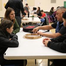 Knowledge Keeper Darren Charlie sits at a table with 2 adults and 1 grade 5 student to demonstrate how to tie the hide to the base of the drum