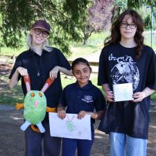 Two female secondary students stand with female elementary student holding their projects