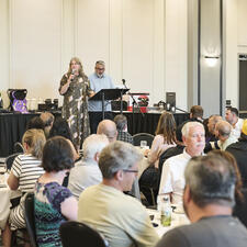 Room full of people sitting at tables with two people standing at a podium speaking into a microphone